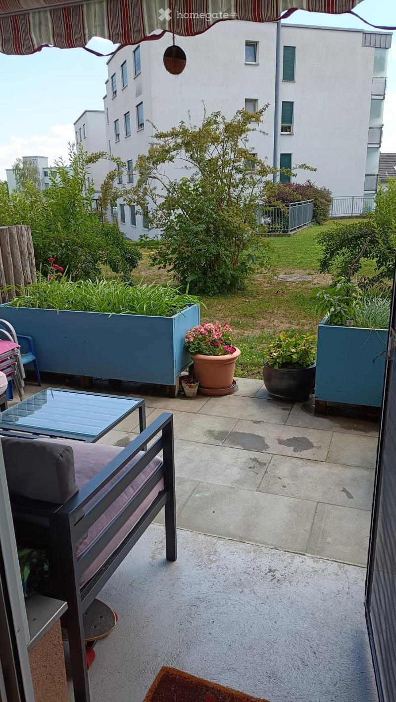 Balcony with a sofa, glass table, chairs, and potted plants, overlooking a landscaped garden area with additional greenery and buildings in the background.