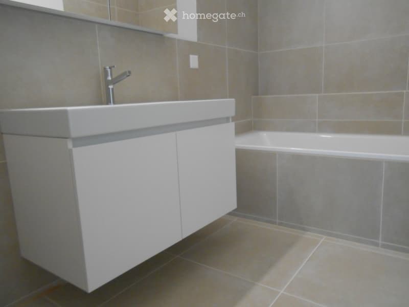 Modern bathroom with a white sink, chrome faucet, and bathtub, featuring grey tiled walls and a beige tiled floor.
