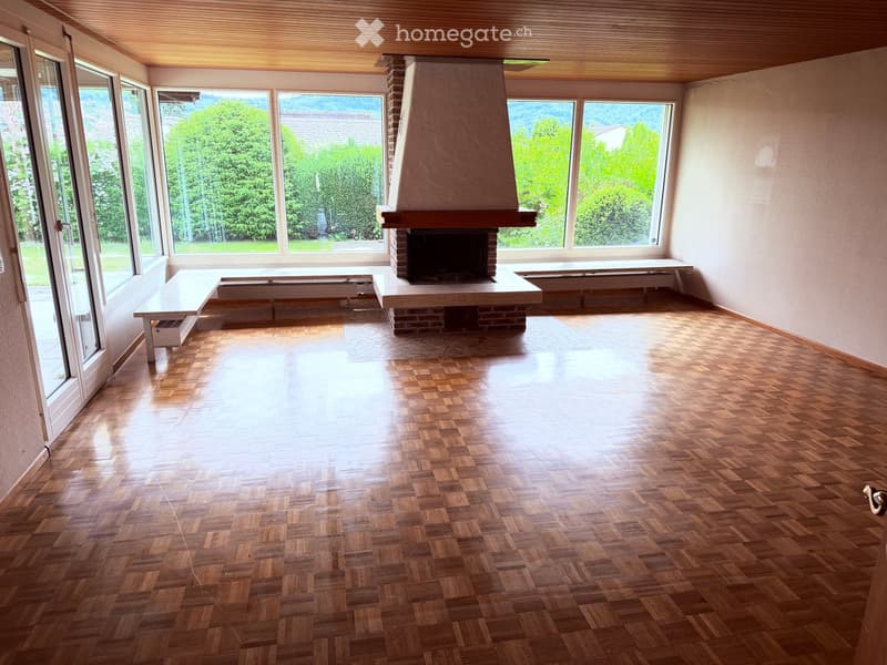 Empty room with wooden herringbone flooring, large windows offering a scenic view, and a central brick fireplace with a wooden mantel.