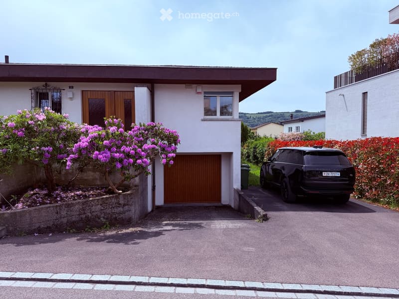 Two-story house with a modern design, featuring a garage, a driveway, and well-maintained landscaping with flowering shrubs and trees.