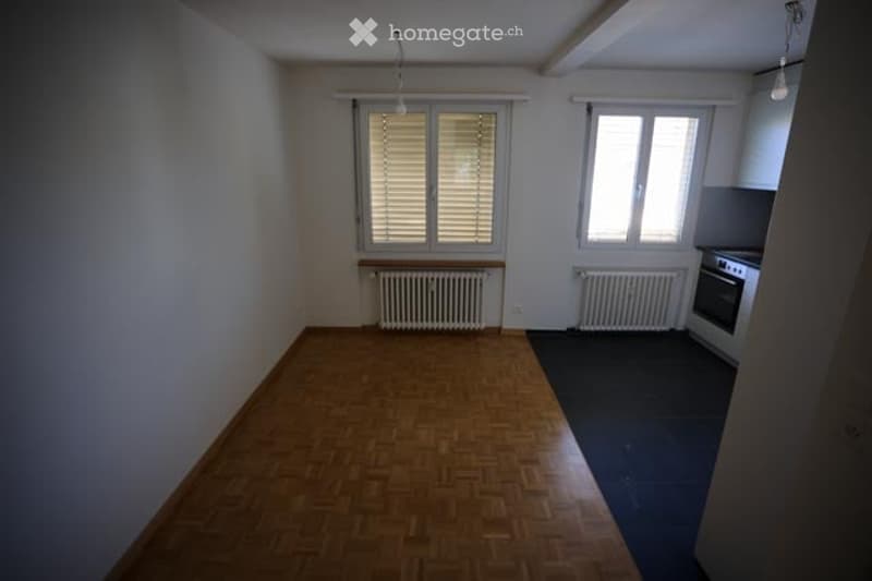Empty room with wooden flooring, white walls, two windows with blinds, radiators, and a small kitchen area with a stove.