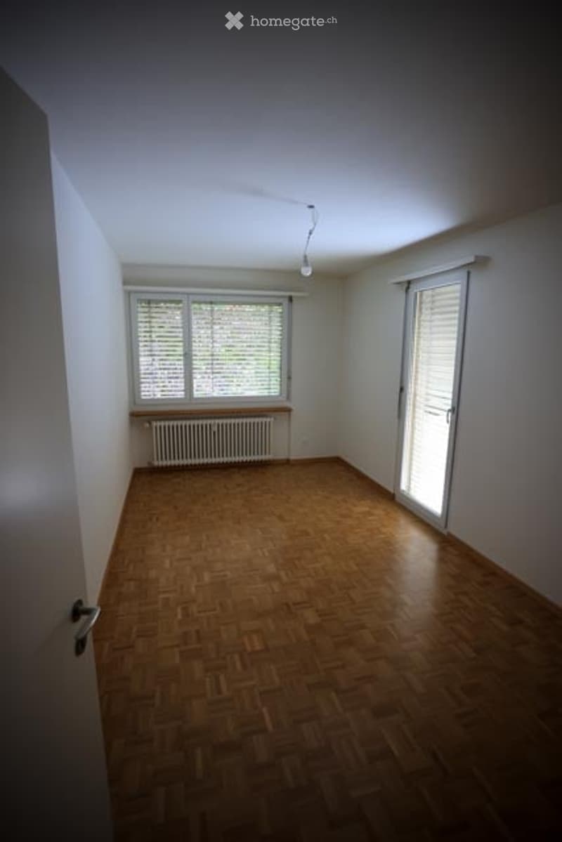 Empty room with wooden flooring, white walls, large windows with blinds, radiator, and a single door.