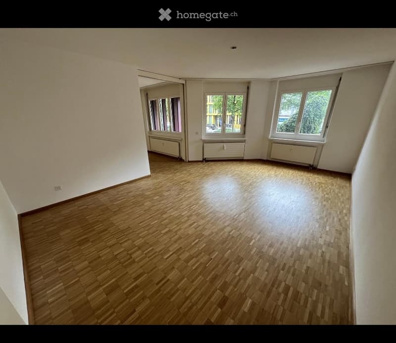 Empty room with wooden flooring, white walls, and large windows with radiators beneath them.