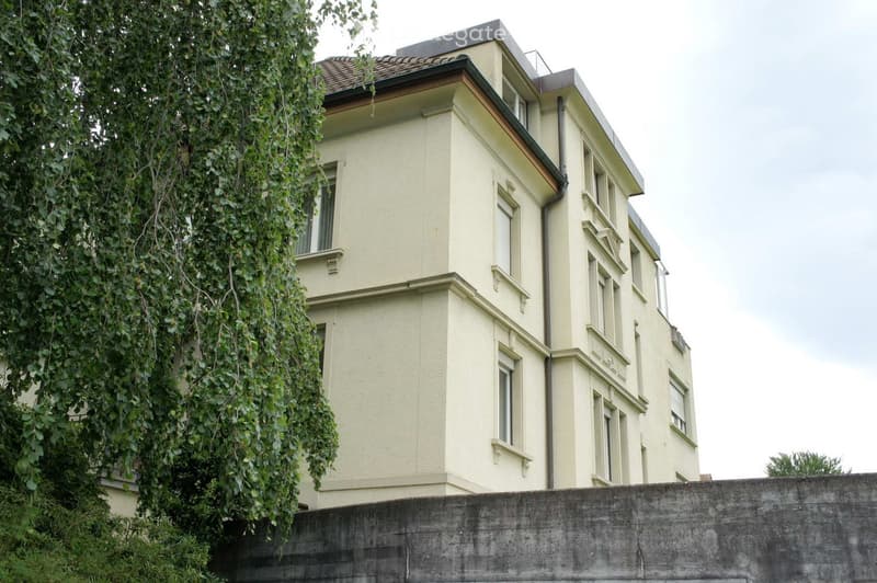A multi-story residential building with light yellow walls, multiple windows, and a tiled roof, partially obscured by a large tree on the left side.