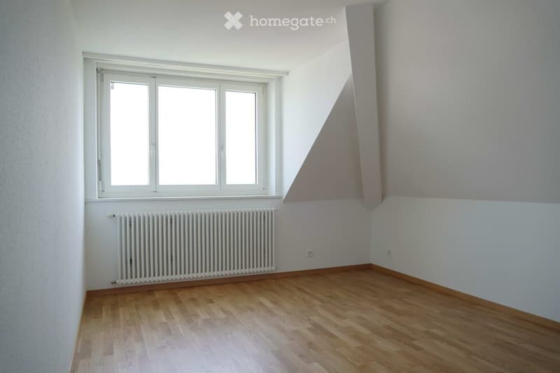 Empty room with wooden flooring, white walls, large window, radiator, and electrical outlets.