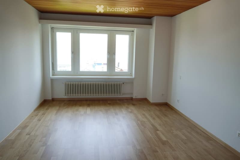 Empty room with light wooden flooring, white walls, large windows, and a radiator below the windows.