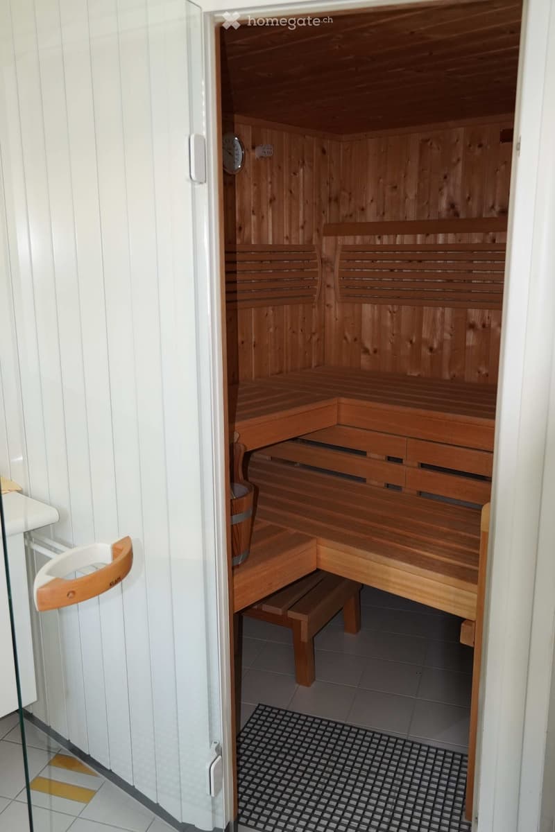 Wooden sauna with multiple levels of benches, steps, and a small wooden bucket, adjacent to a tiled bathroom area with a glass door.