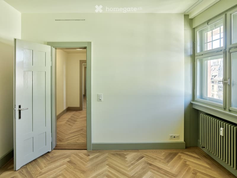 Empty hallway with light-colored walls, wooden herringbone flooring, and a white door leading to another room. Windows on the right side provide natural light.