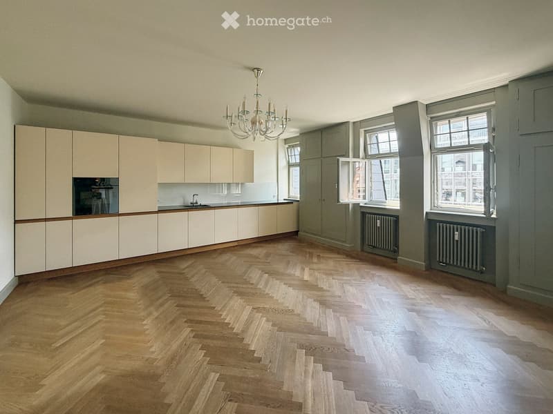 Empty kitchen with light-colored cabinets, wooden herringbone flooring, and large windows. No appliances or furniture are present.