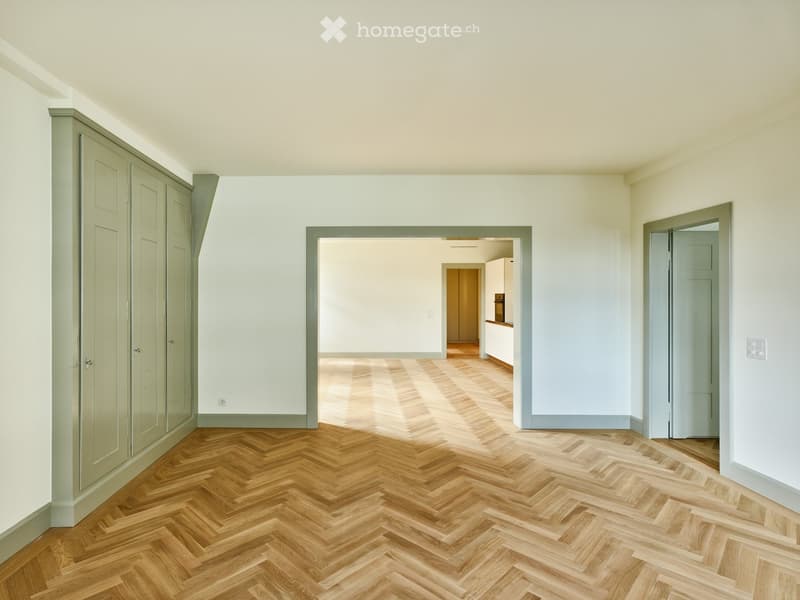 Empty room with herringbone wooden flooring, white walls, and grey-framed doors leading to other rooms.