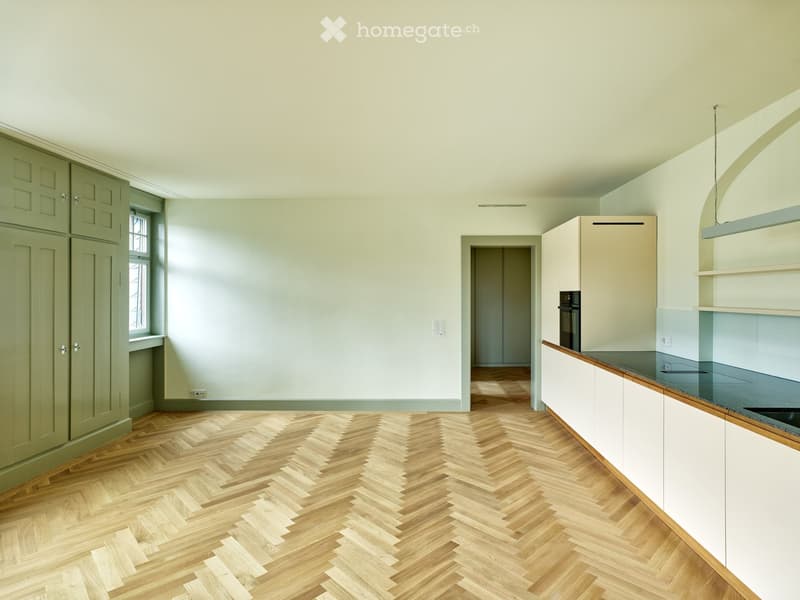 Empty kitchen with light green walls, herringbone wooden flooring, white cabinets, and a dark countertop. No appliances or furniture are present.