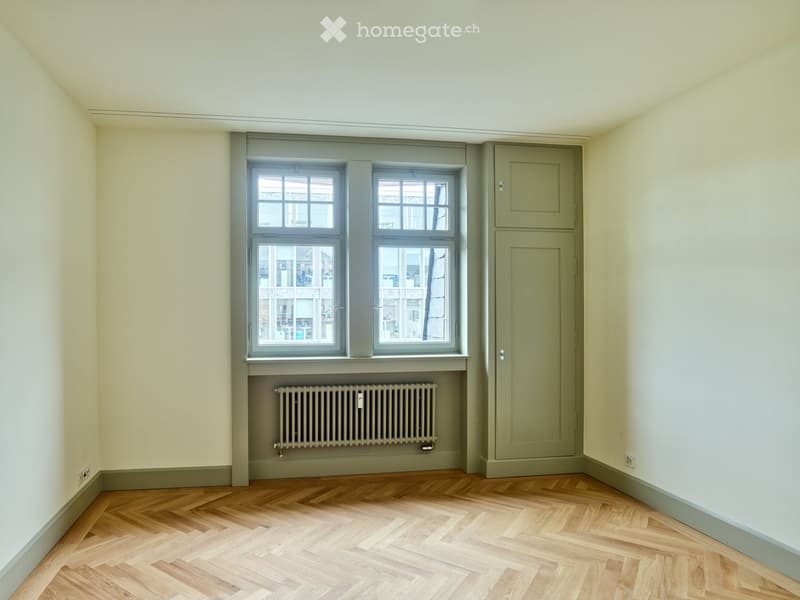 Empty room with light-colored walls, wooden herringbone flooring, and a radiator beneath a double window. The room has a simple, uncluttered design with no furniture or personal items.