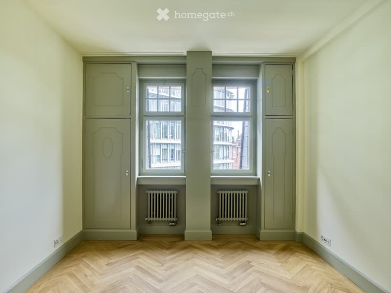 Empty room with light green painted walls, wooden herringbone flooring, two windows with radiators below, and built-in cabinets on either side.