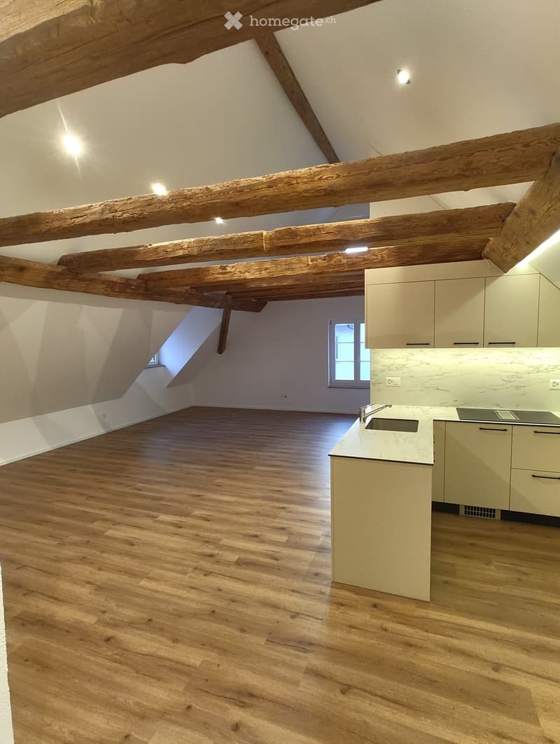 Empty attic space with wooden beams, wooden flooring, and a small kitchen area with cabinets and a sink.