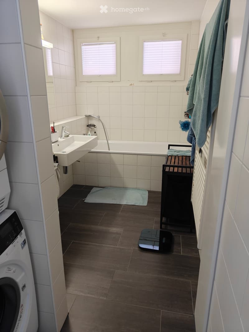 Bathroom with white tiled walls, a sink, bathtub, washing machine, a small wooden stool, and towels hanging on the wall.