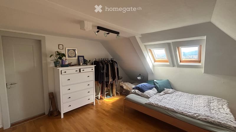 Cozy attic bedroom with wooden floors, slanted ceilings, and large windows offering a view. Features a bed with pillows and blankets, a dresser, a clothing rack, and a small plant on the dresser.
