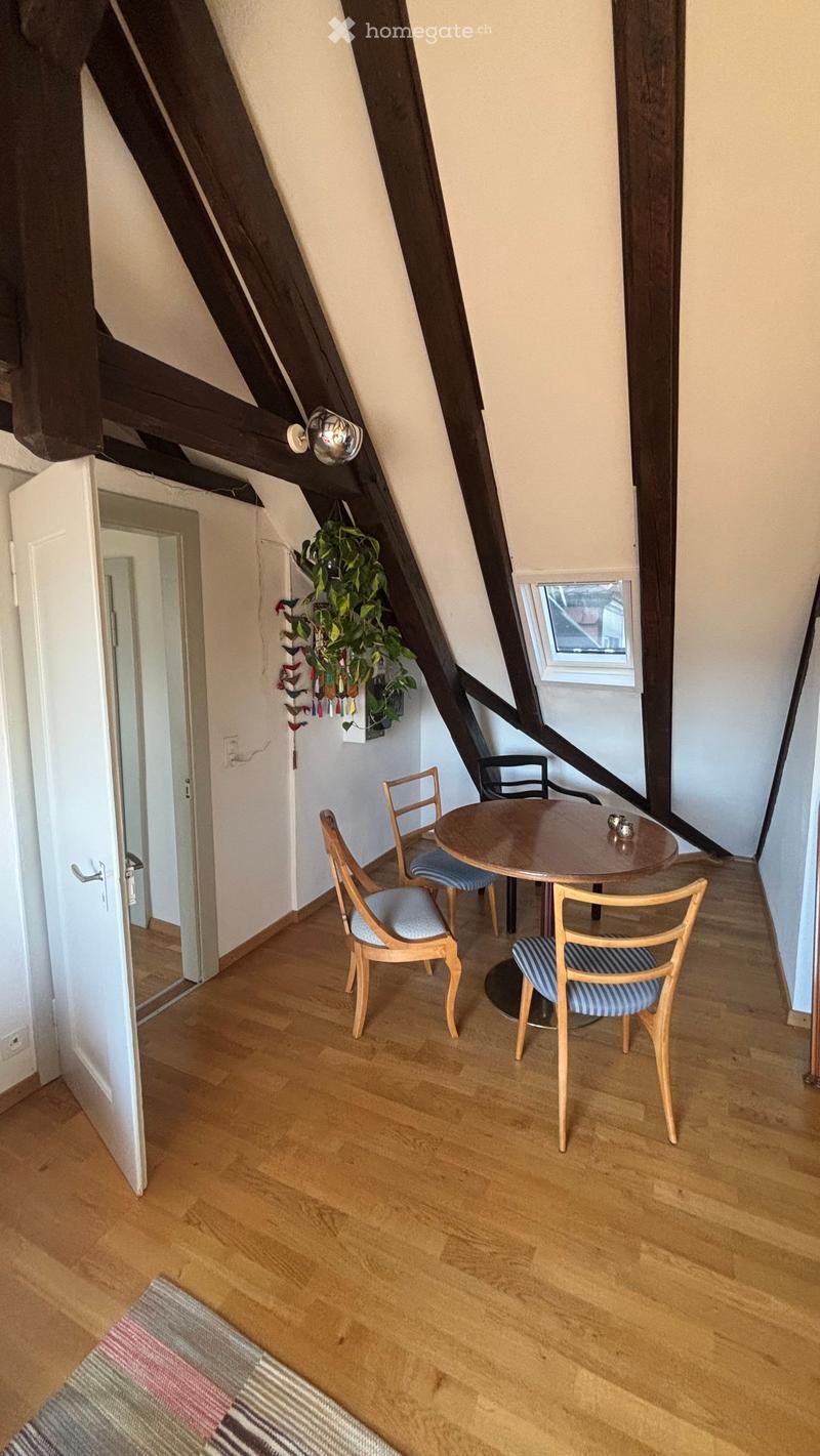 Cozy attic room with wooden floors, slanted wooden beams, a small round table with four chairs, a hanging plant, and a colorful rug on the floor.