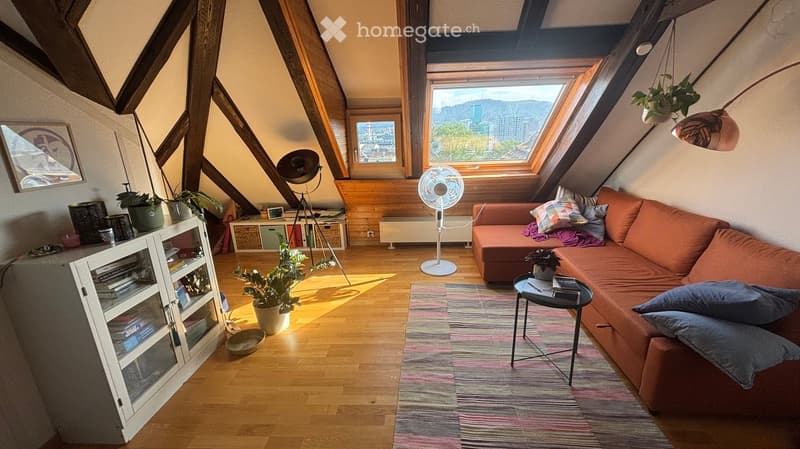 Living room with wooden floor, slanted ceiling, large windows offering a city view, orange sofa, coffee table, cabinet, fan, floor lamp, and potted plants.