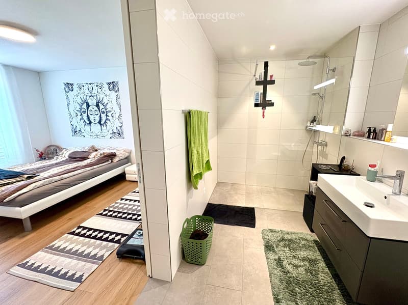 The image shows a modern, compact bedroom and bathroom combination. The bedroom features a neatly made bed with pillows and blankets, a patterned rug, and a wall-mounted artwork. The adjacent bathroom includes a sink with a modern faucet, a shower with a