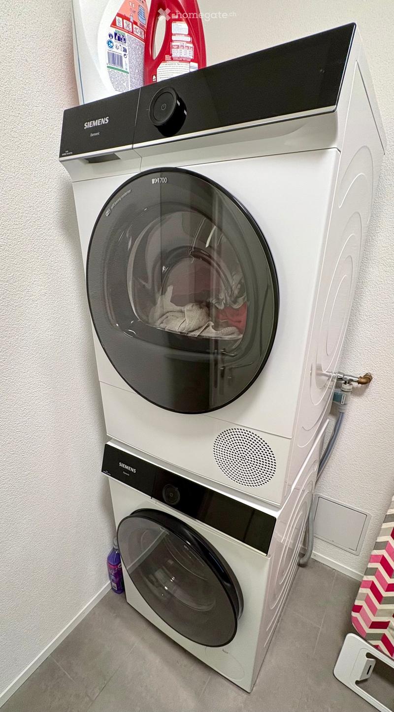 A modern bathroom with a white Siemens washing machine and a dishwasher stacked vertically, with cleaning supplies visible on top.