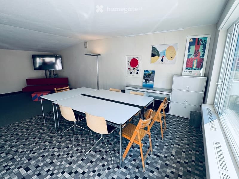Bright room with tiled floor, large white table surrounded by chairs, red sofa, filing cabinet, and several artworks on the walls.