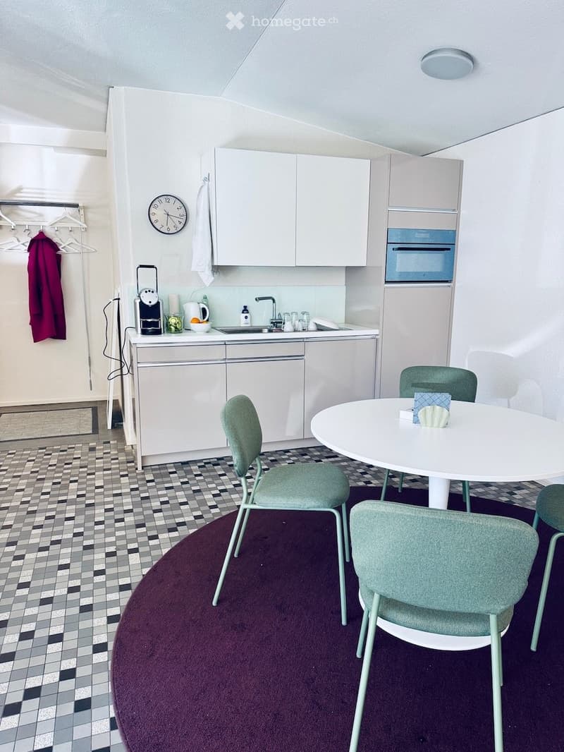 A modern kitchen and dining area with white cabinets, a round dining table with four chairs, a coffee maker, and a clock on the wall.