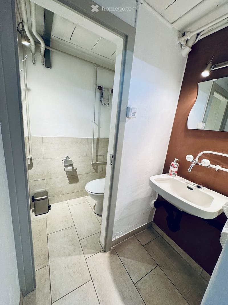 Modern bathroom with white tiled walls and floor, a toilet, a sink with a modern faucet, a mirror, a soap dispenser, and a towel hanging on the wall.