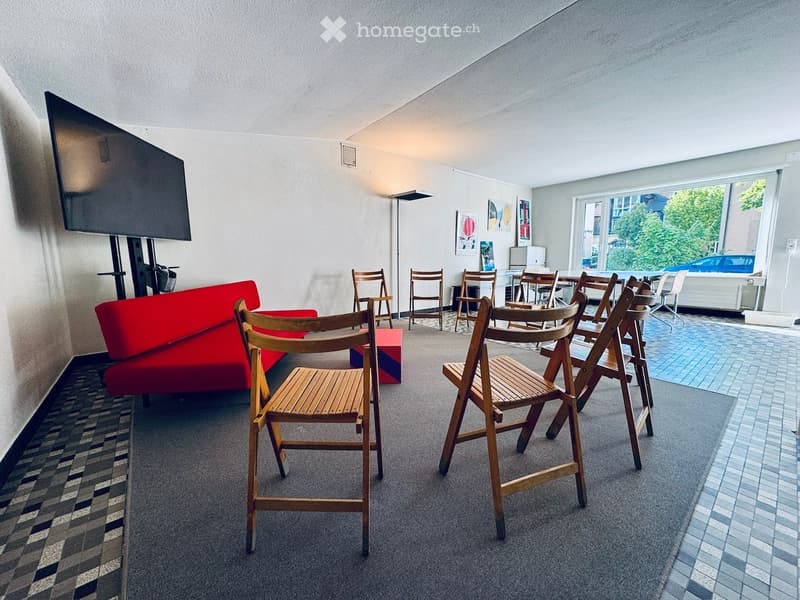 Modern, open-plan room with a red sofa, multiple wooden chairs, a small table, a large screen mounted on the wall, and a lamp. Large windows provide natural light and a view of greenery outside.