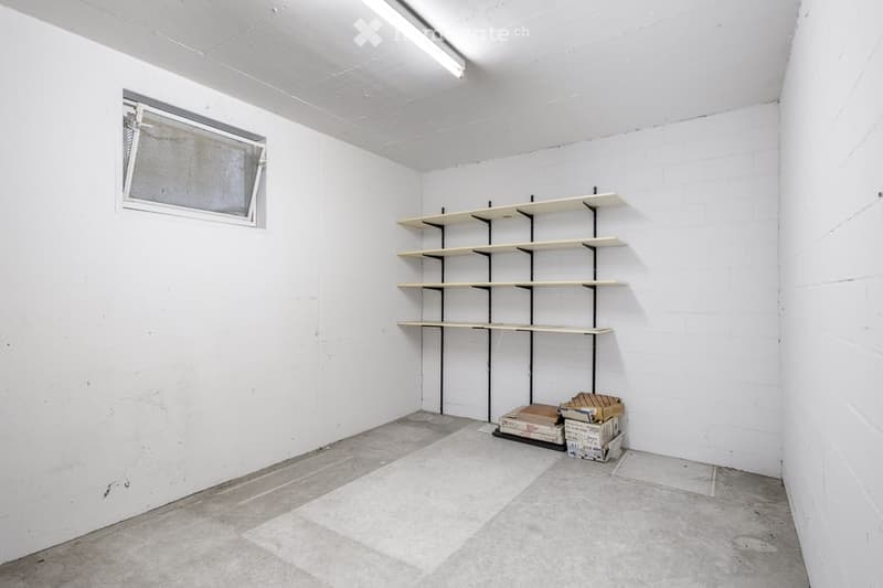Empty room with white walls, concrete floor, and a small window. Includes a set of empty shelves and some stacked boxes in the corner.
