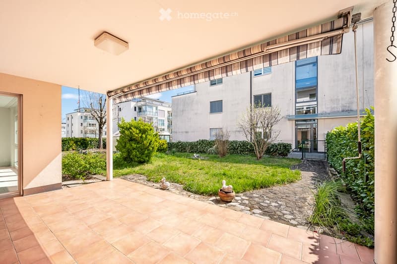 A well-maintained garden area with a tiled patio, small fountain, and surrounding greenery, featuring a view of neighboring buildings.