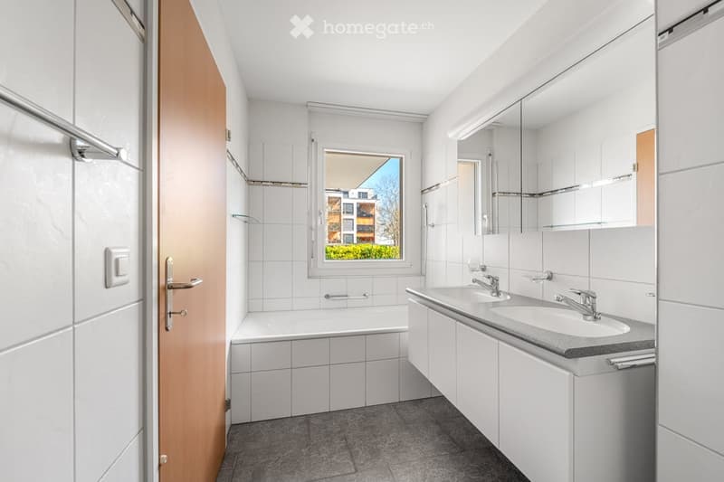 Modern bathroom with white tiles, a bathtub, a double sink, and a large mirror. A window offers a view of the outside.