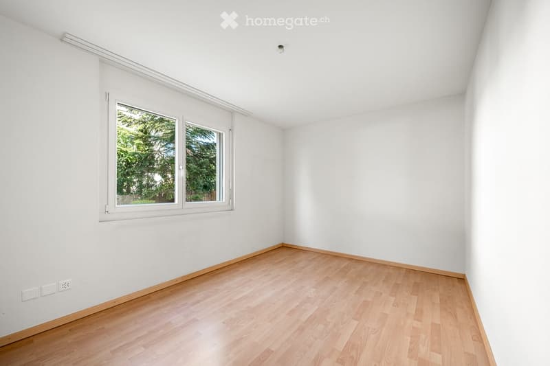 Empty room with white walls, wooden flooring, and large windows showing greenery outside.