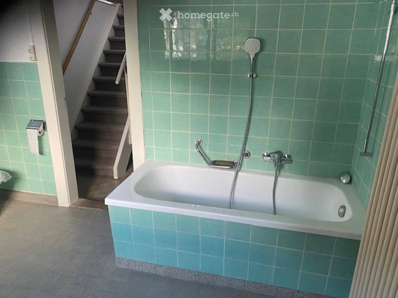 Bathroom with a white bathtub, green tiled walls, a showerhead, and a handrail for support.