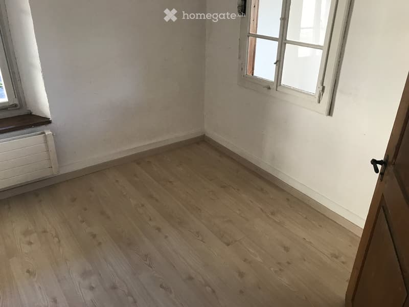 Empty room with wooden flooring, white walls, and two windows with white frames. A radiator is visible under one window, and a door is partially open on the right side.