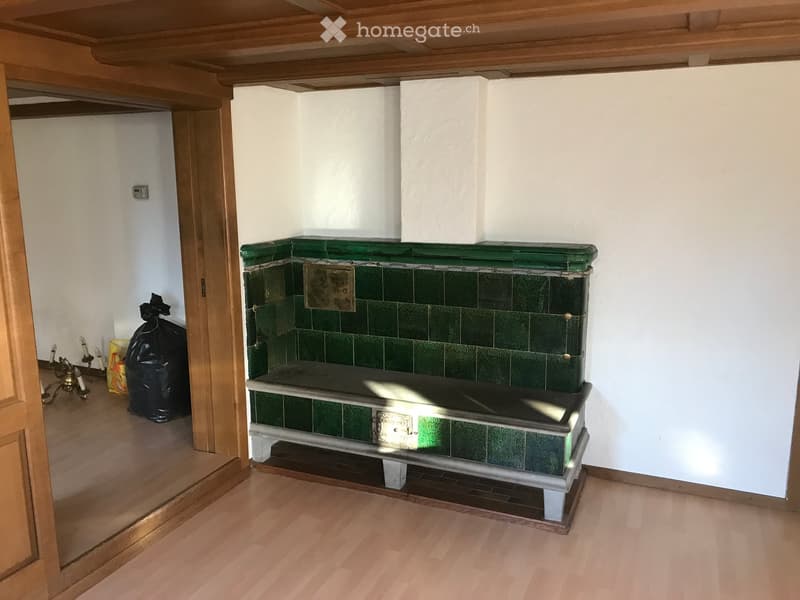 An unfurnished room with a fireplace, wooden floor, and white walls. The fireplace has green tiles and a decorative design.