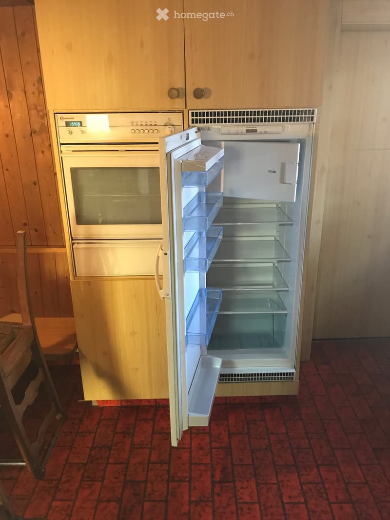 A kitchen with a red tiled floor, wooden cabinets, and an open refrigerator next to an oven.