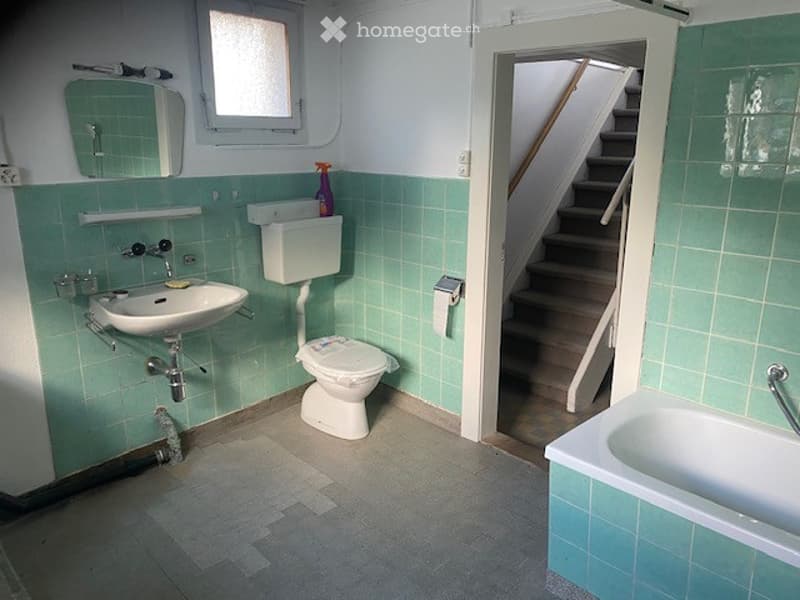Bathroom with green tiled walls, a sink, toilet, bathtub, and a mirror above the sink. A small window is visible above the sink, and a doorway leads to a staircase.