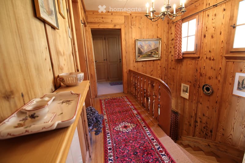 Wooden-paneled hallway with a chandelier, framed artwork, a red runner, and a small table with decorative items.