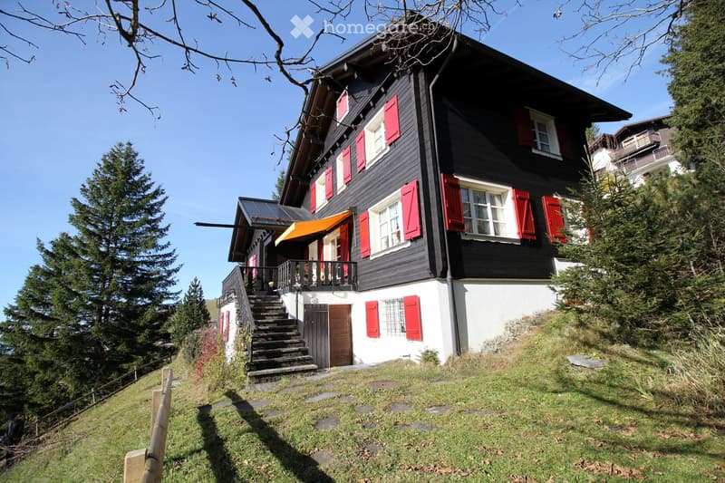 A two-story house with black exterior walls, red shutters, and white trim. Features include a staircase leading to a balcony, a covered entrance, and a solar panel on the roof.
