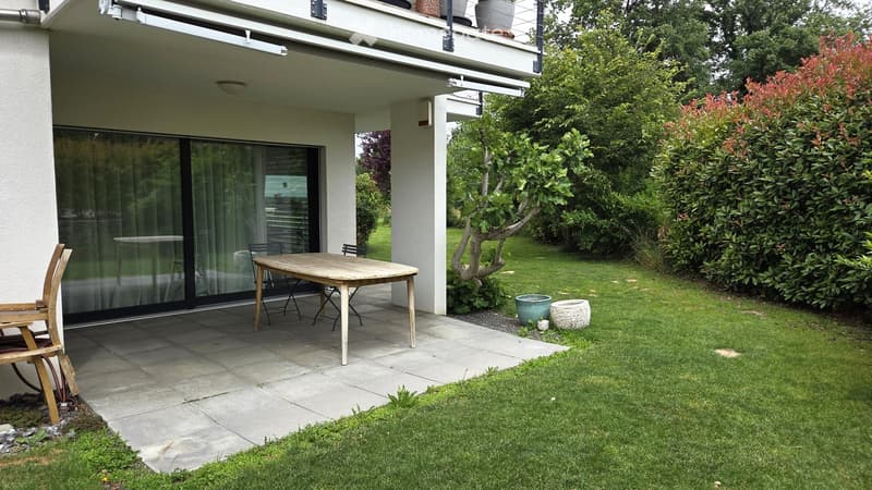 Garden area with a wooden table and chairs, surrounded by greenery and a modern house with a balcony.