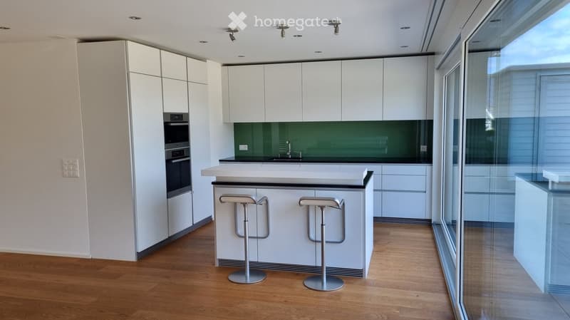 Modern kitchen with white cabinets, a central island, and two stools, featuring a green backsplash and large windows.