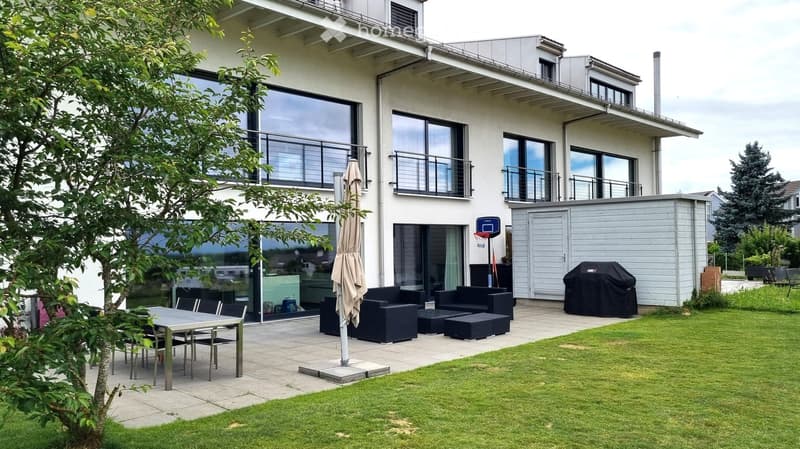 Modern outdoor patio area with a dining table, chairs, black sofa, ottoman, and a covered grill. The patio is adjacent to a two-story house with large windows and balconies.