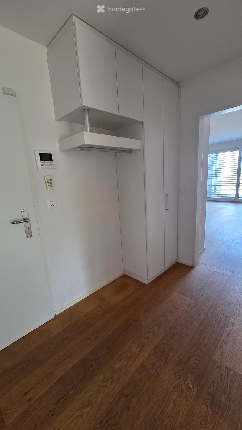 Empty room with light wooden flooring, white walls, built-in white cabinets, and a window with blinds leading to a balcony.