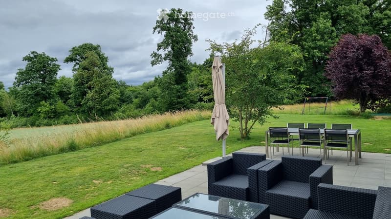 Outdoor patio with wicker furniture, including a table, chairs, and an umbrella, surrounded by a well-maintained lawn and trees.