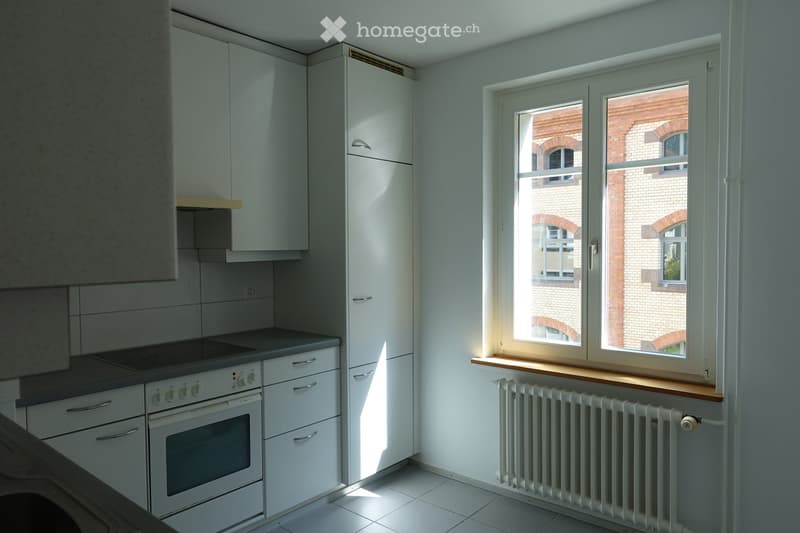 Modern kitchen with white cabinets, a built-in oven, and a radiator beneath a large window overlooking a brick building.