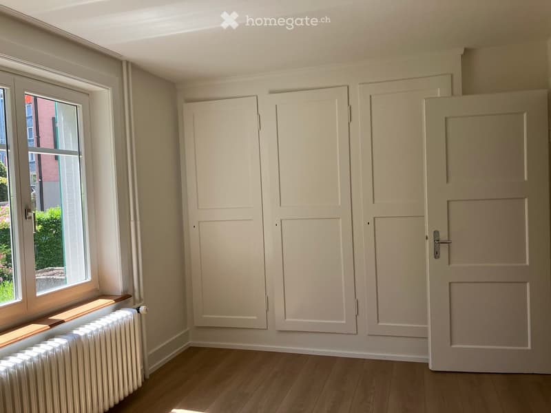 Empty room with light-colored walls, wooden flooring, and multiple doors. A window with a radiator below it is visible on the left side.