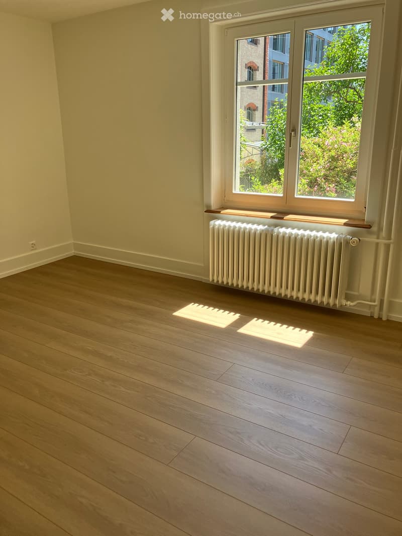 Empty room with light wooden flooring, white walls, and a window with a radiator below it. Sunlight streams in from the window, casting shadows on the floor.