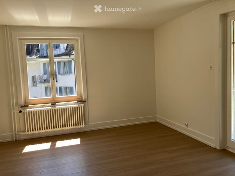 Empty room with wooden flooring, white walls, large window with view of neighboring building, radiator below window, and natural light streaming in.
