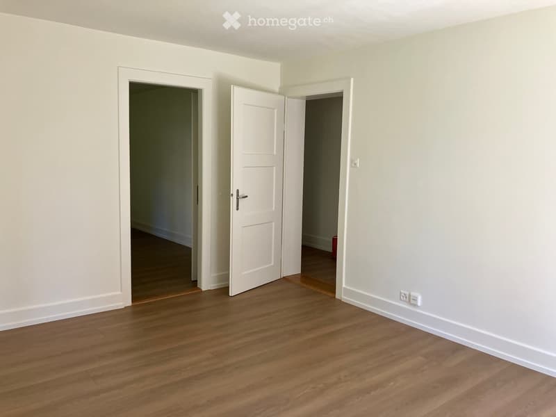 Empty room with light-colored walls and wooden flooring, featuring two open doors leading to adjacent spaces.