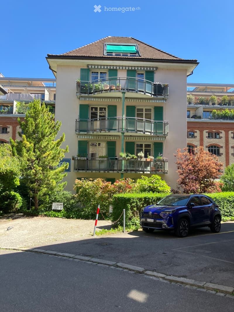 A multi-story residential building with green shutters, balconies, and potted plants. A blue car is parked in front.